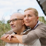 Eine Frau umarmt ihren Mann vor einem Holzhaus, während sie in die Ferne sehen